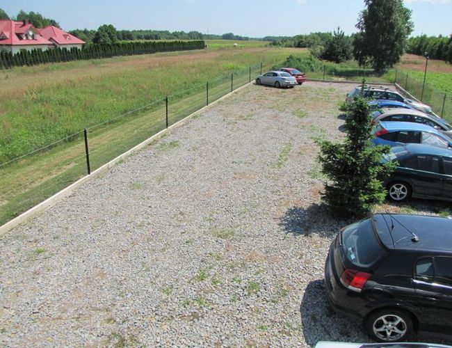 Parking Auto Park - Ogrodzony plac z żwirową nawierzchnią. Kilka samochodów stoi po bokach. W tle trawniki i pola.