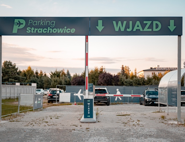 Parking Strachowice – Widoczne jest wejście z szlabanem. Teren otoczony jest ogrodzeniem. Na parkingu stoją samochody.