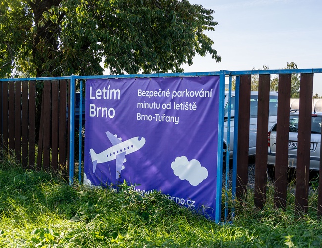 Parking Letim Brno – Na ogrodzeniu widoczny jest duży baner. Obok stoją zaparkowane samochody. W tle rośnie drzewo.