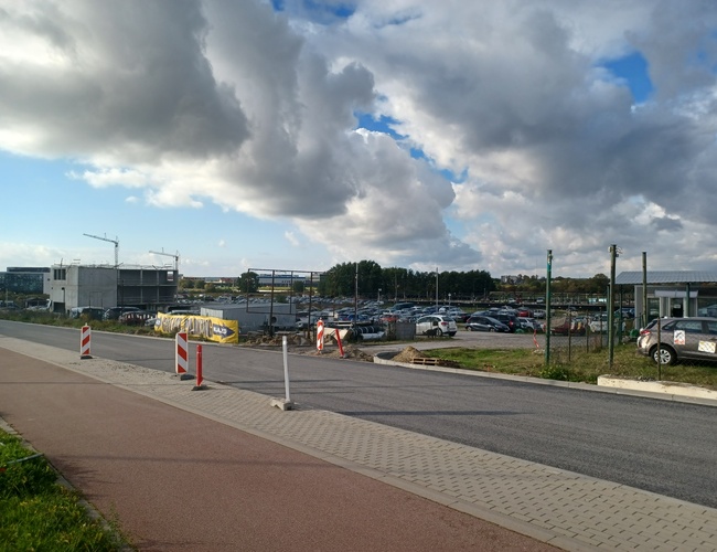 Parking Goliat – Duży, ogrodzony teren z wieloma samochodami. W tle widać budowę. Na niebie unoszą się ciemne chmury.