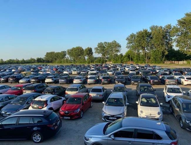 Smart Premium Parking - Gran estacionamiento al aire libre, lleno. Muchos coches están estacionados cerca unos de otros en filas. Los árboles al fondo rodean el área.