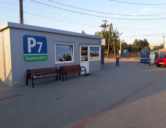 Parking P7 – Budynek z oknami i ławkami z przodu. Obok stoi czerwony samochód. Teren jest ogrodzony.