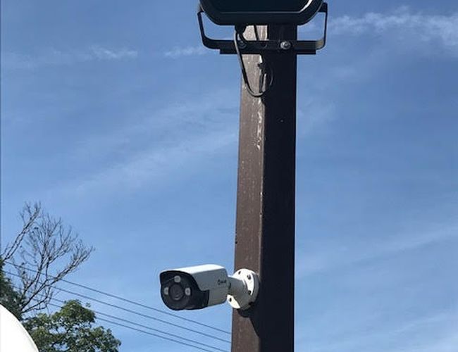 Auto Airport Samoobsługowy – Na słupie znajdują się kamera i lampa. W tle widać niebo oraz drzewa.