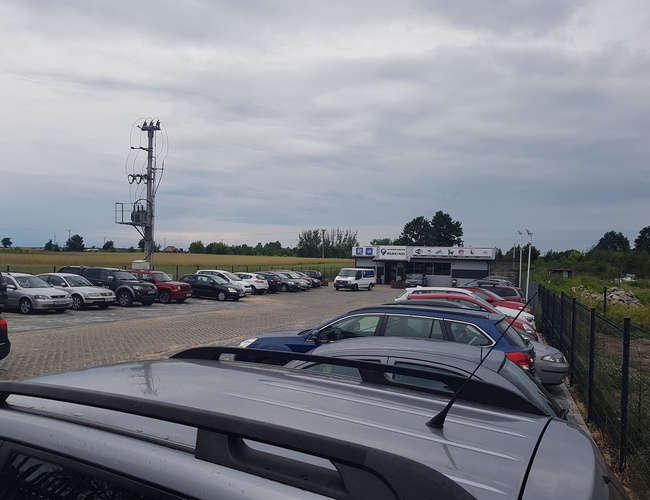 PARKING P24 PREMIUM - Parking pełen samochodów na utwardzonej nawierzchni. Teren jest ogrodzony i oświetlony. W tle stoi budynek z oznaczeniami.