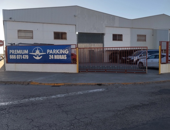 Premium Parking VLC - Budynek z metalowym ogrodzeniem i bramą. Znajduje się tam kilka zaparkowanych samochodów. Na ogrodzeniu widnieje reklama z godzinami otwarcia.
