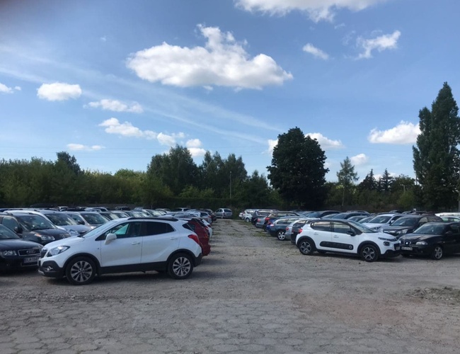 Parkuj Leć – Parking pełen samochodów na otwartym terenie. W tle drzewa i niebo z chmurami. Teren jest nieutwardzony.