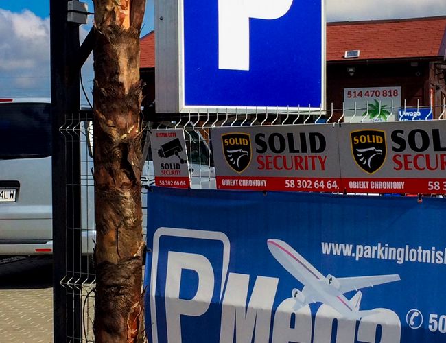 Parking Mega Park Service – Znak z literą P na niebieskim tle. Ogrodzenie zabezpiecza parking. Reklamy zabezpieczeń na płocie.