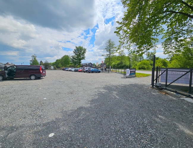 ParkZone – Štrkový parkovací priestor s oplotením a stromami. Zaparkované autá. Čiastočne zamračená obloha s prestávkami.