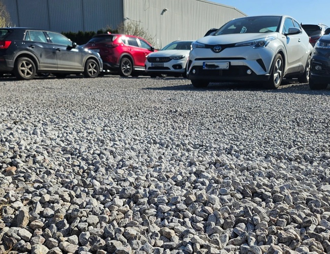 Just Fly Parking – Żwirowy plac z zaparkowanymi samochodami. Teren otwarty z metalowym budynkiem w tle.