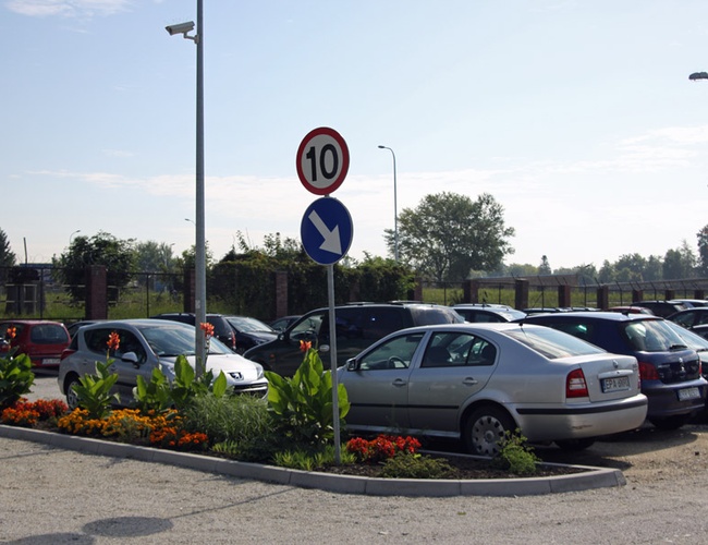 Parking Rondo – Na zdjęciu są samochody i kwiaty. Widoczne jest także ogrodzenie oraz znaki drogowe. Cały teren jest dobrze oświetlony.