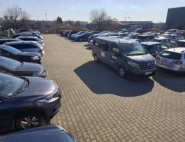 Parkovisko Olo Car Park - Na parkovisku stojí niekoľko osobných áut a dodávok. Povrch je vydláždený kockami. Na pozadí rastú stromy.