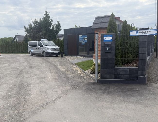 FlyParking – Przy wjeździe stoi auto transportowe. Teren jest ogrodzony metalowym płotem i otoczony zielenią.