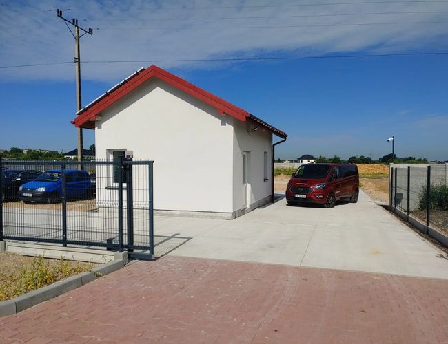 Estacionamiento de toros - puerta de entrada al lado del pequeño edificio con techo rojo. El estacionamiento está cercado y pavimentado. Hay un coche rojo allí.