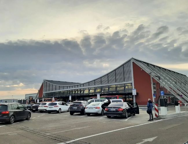 Parking z zaparkowanymi samochodami przed nowoczesnym budynkiem o przeszklonej elewacji. Przed wejściem widoczne są osoby poruszające się po terenie obiektu.