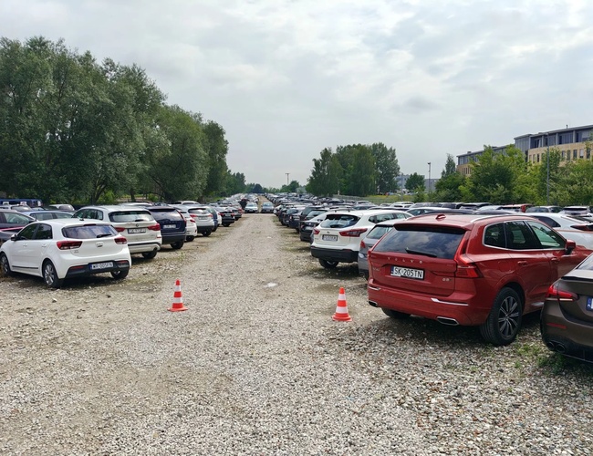 WAW Parking – Poarta de acces cu banner-e publicitare. Suprafață de pietriș înconjurată de un gard. Copaci în fundal.
