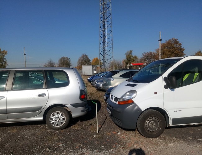 Parking Bastion - Los coches están en un área de grava. El terreno está abierto y sin cercas. Al fondo se ven árboles y postes.