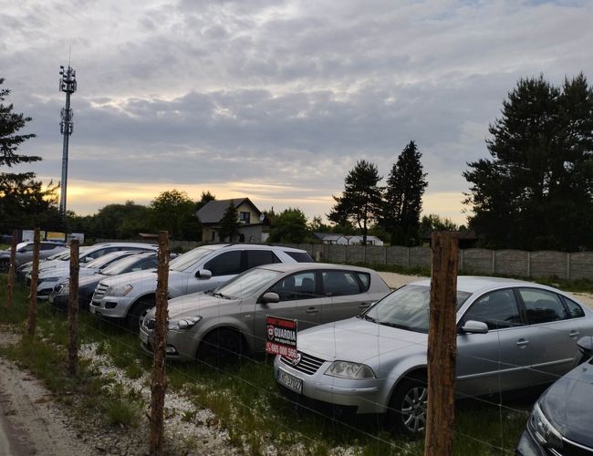Parkplatz Guardia – Eine Reihe von Autos steht neben dem Zaun aus Maschendraht. Das Gelände ist von großen Bäumen und einer Betonmauer umgeben.