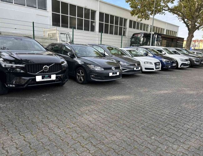 Parking Parkinsel24 VALET - Rząd samochodów stoi obok budynku. Parking jest ogrodzony metalowym płotem. W tle widać jeden drzewo.