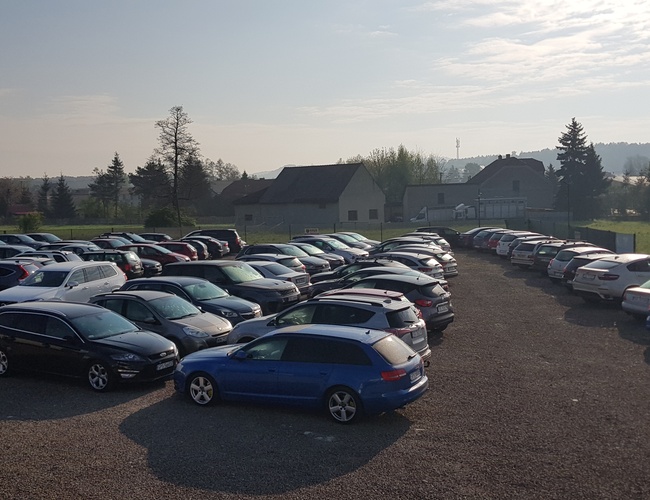 BaliceLot24 – Je viditeľné veľké, nezpevnené parkovisko s viacerými autami. Pozemok je ohraničený plotom a okolie zelene dodáva otvorenosť.