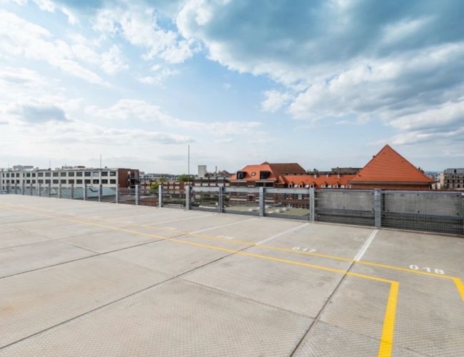 Parking Best Valet Park Service - widoczny jest betonowy dach parkingu. Na tle nieba widać budynki z czerwonymi dachami. Krawędź parkingu jest ogrodzona.