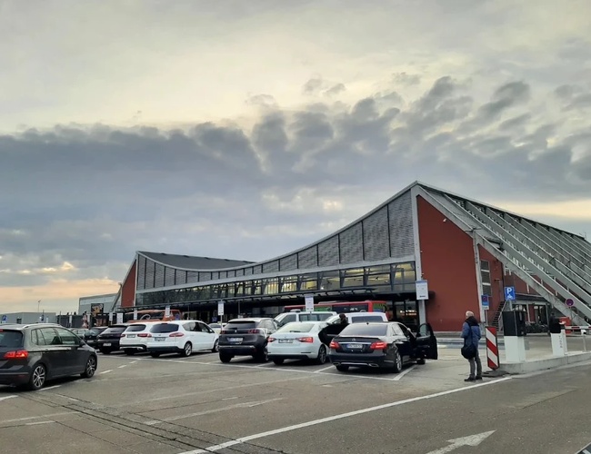 Parking z zaparkowanymi samochodami przed nowoczesnym budynkiem o przeszklonej elewacji. Przed wejściem widoczne są osoby poruszające się po terenie obiektu.