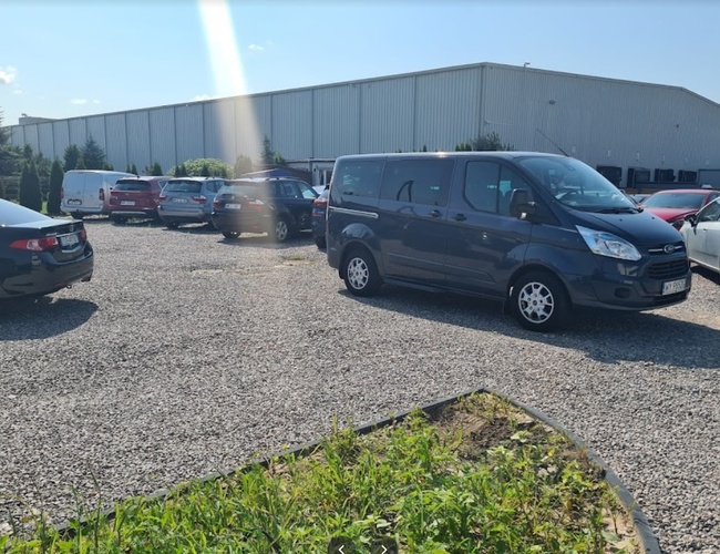 Parkovanie Dominik - Na kamenistej ploche stoja autá. Pozemok je nezastrešený a obklopený zeleňou. V pozadí je vidieť veľkú budovu.