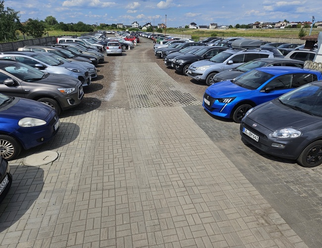 Estacionamiento U Madzi - Filas de coches estacionados a lo largo del camino de piedra. El área está cercada y iluminada.