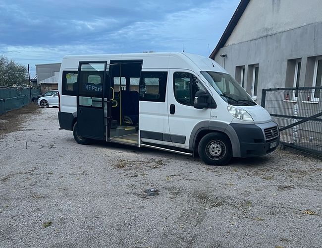 Biały bus zaparkowany na żwirowym placu obok budynku, drzwi pojazdu są otwarte