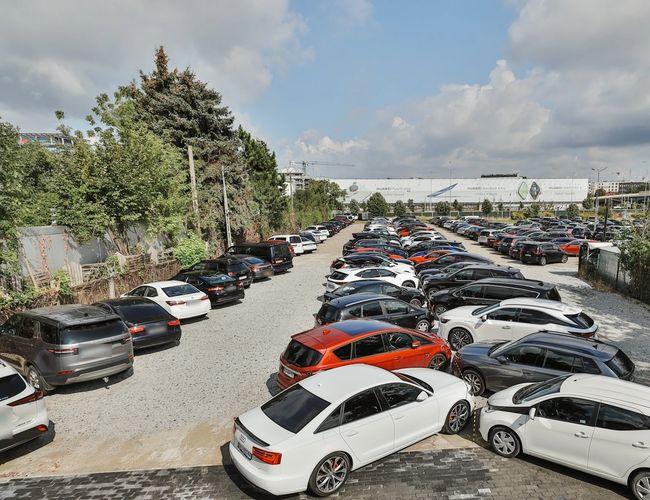 Smart Supreme Parking – na żwirowym placu stoją zaparkowane auta. Otaczają go drzewa i ogrodzenie. Niebo jest pochmurne.
