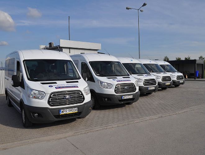 Parking Lotnisko – Białe busy stoją w rzędzie. Teren jest asfaltowy i przestronny. W tle znajduje się zadaszenie.