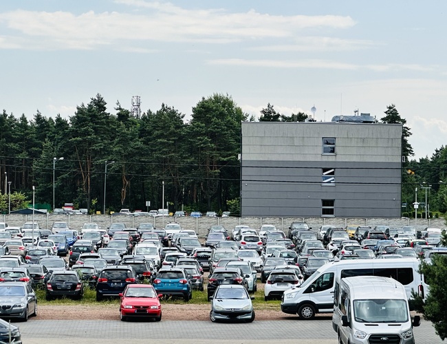 Comienza con nosotros - El aparcamiento está lleno de coches. Al fondo se ven árboles y un edificio. Las luces iluminan el área.
