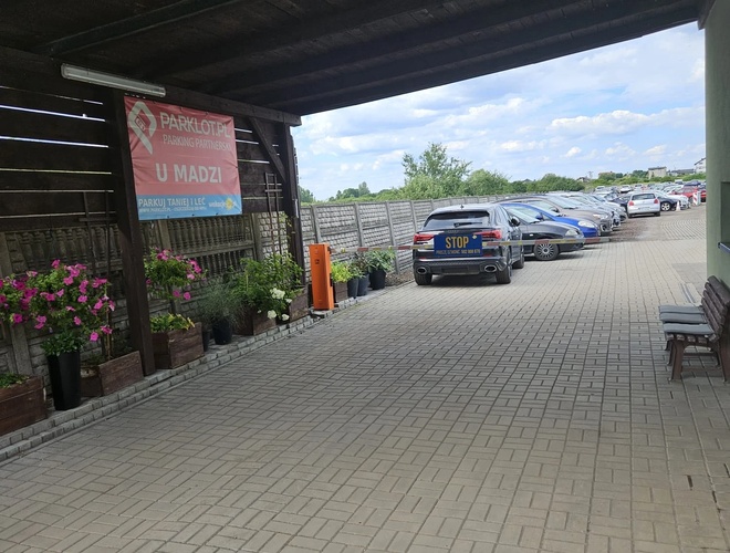 Aparcamiento U Madzi – Entrada cubierta, superficie pavimentada. Flores en la entrada, coches aparcados en fila.