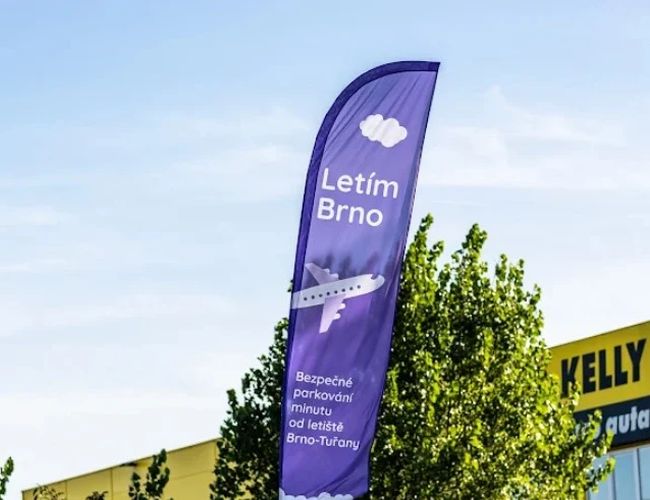 Parking Letim Brno II – na zdjęciu widoczna jest fioletowa flaga reklamowa. Znajdują się na niej napisy informacyjne oraz grafika samolotu. W tle widać drzewo i fragment budynku.