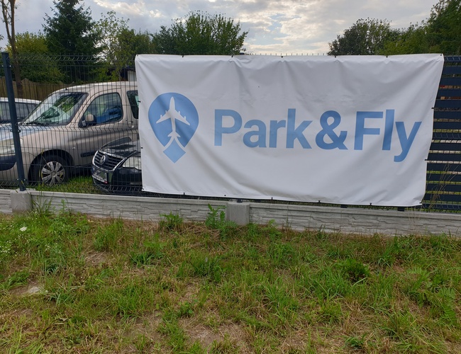 Parkovanie Park&Fly - oplotené územie s zaparkovanými autami. Na oplotení je veľký billboard.