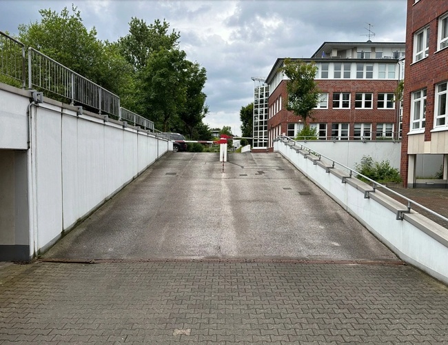 Parking Marspark Outdoor Valet – Wjazd podjazdem ograniczonym barierką. Widoczne drzewo i budynek z czerwoną cegłą. Niebo jest pochmurne.