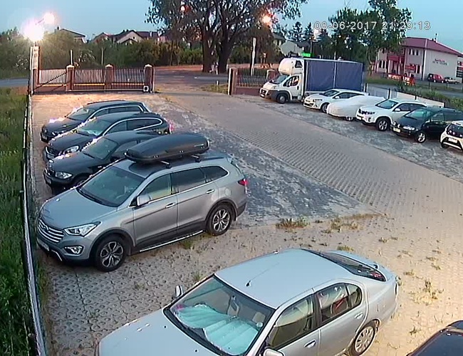 PARKING P24 PREMIUM – Parking jest ogrodzony i oświetlony. Kilka aut zaparkowanych obok siebie. W tle widać bramę i budynki.