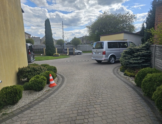 Estacionamiento en Krótkiej – área pavimentada con arbustos. Lugares disponibles para coches. Al fondo, una cerca y una piedra decorativa.