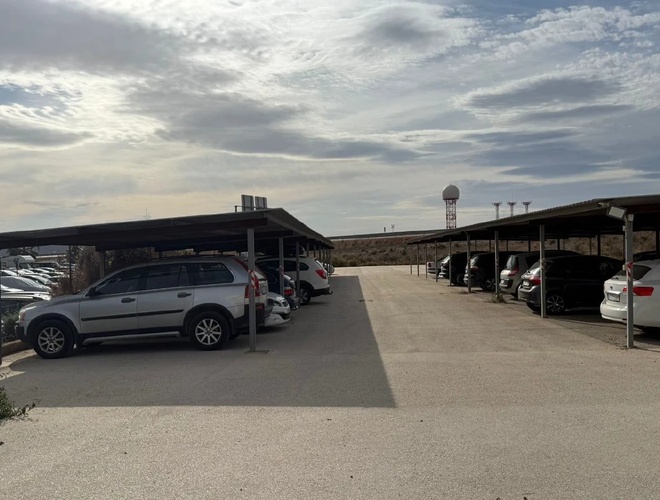 Szeroki parking z kilkoma zaparkowanymi autami pod zadaszonymi miejscami, w tle więcej zaparkowanych samochodów pod zadaszeniem i na utwardzonym terenie.