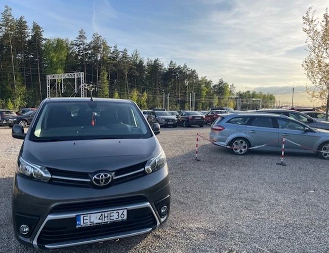 Auto Airport Parking – Na parkingu stoją zaparkowane samochody. Teren jest utwardzony i częściowo ogrodzony. W tle widać drzewa.