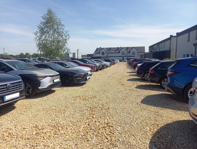 AutoParking24h - Żwirowy plac wypełniony samochodami stojącymi w równych rzędach. W tle widać budynki i drzewa.