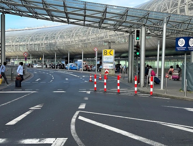 Паркування Marspark Outdoor Valet – на знімку зображено оживлену дорогу біля аеропорту. Видно накритий тротуар і парковочні розмітки. Люди з валізами йдуть у напрямку входу.