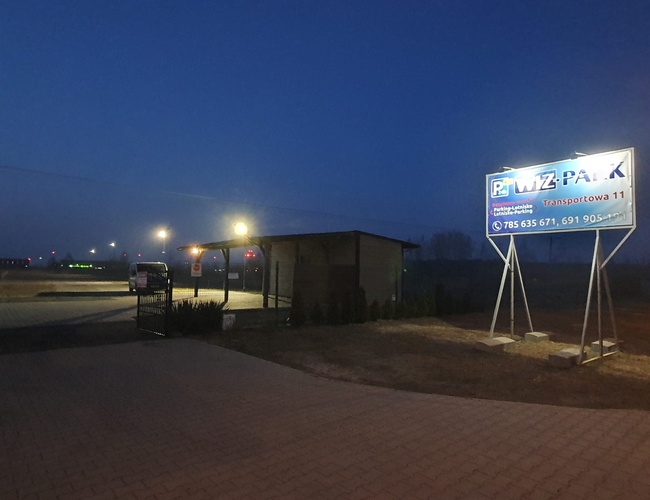 WIZ-PARK – El estacionamiento está iluminado y cercado. Se ve un gran cartel con el nombre. A la izquierda hay un autobús estacionado.