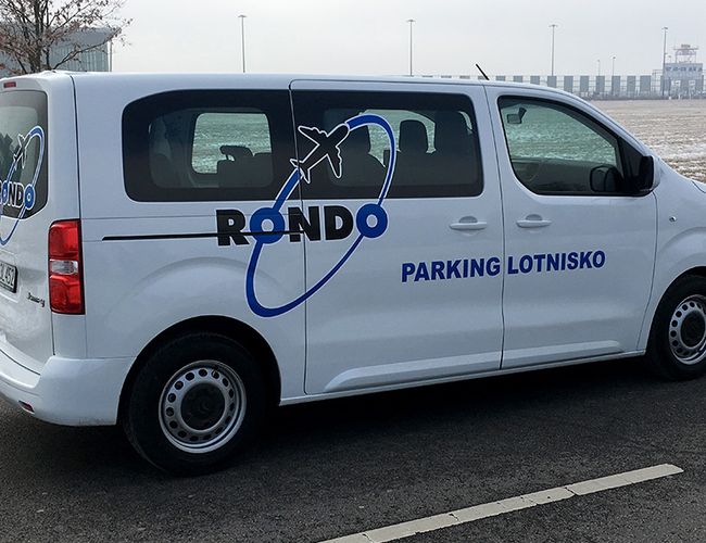 Parking Rondo - Biały van z logo Parking Lotnisko. Zaparkowany na asfaltowej drodze obok lotniska. W tle widoczny teren trawiasty.