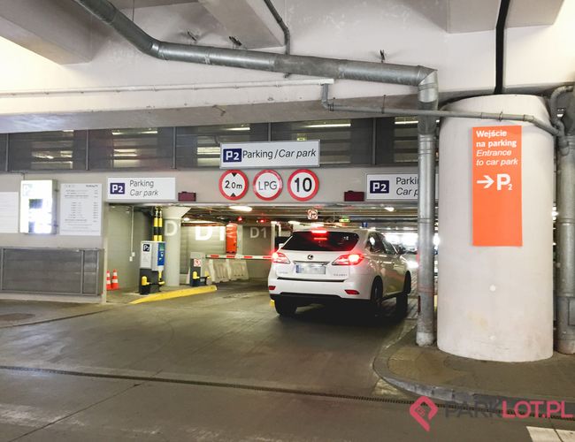 Oficjalny Parking Lotniska P2 – Samochód wjeżdża na zadaszony parking. Jest niski limit wysokości. Znak wskazuje kierunek do wejścia.