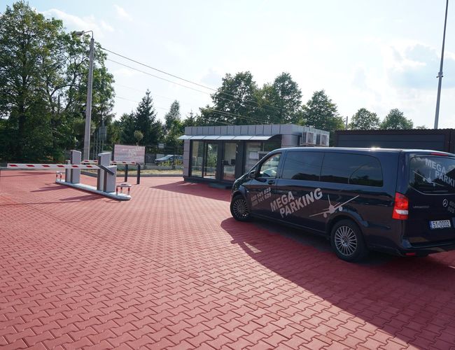 VIP Garaże MEGA Parking - Na parkingu stoją van i barierki. Teren jest ogrodzony. W tle znajdują się drzewa.