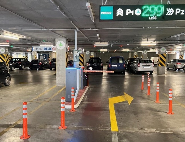 Parkovisko P2 - Okęcie Północ Viacposchodové – priestranné podzemné parkovisko. Sú tu označenia smerov a mnoho zaparkovaných áut.