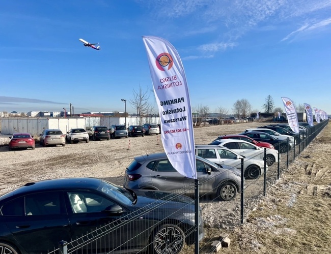 Parkovanie blízko letiska - Automobily stoja na oplotenom námestí. Nad parkoviskom lieta lietadlo. Na plote sú vyvesené vlajky.