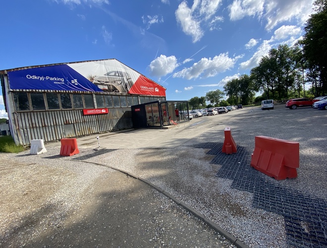 Odkryj-Parking – Duży plac z samochodami przy budynku. Teren częściowo ogrodzony barierkami. Samochody są zaparkowane w rzędach.