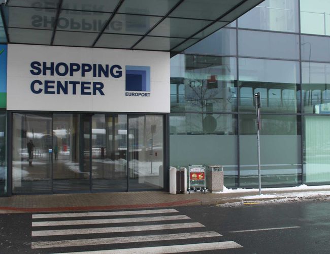 Parking Smile Parking – Wejście do centrum handlowego z przeszkloną fasadą. Przy drzwiach stoją wózki i kosze na śmieci. Na ziemi leży śnieg.