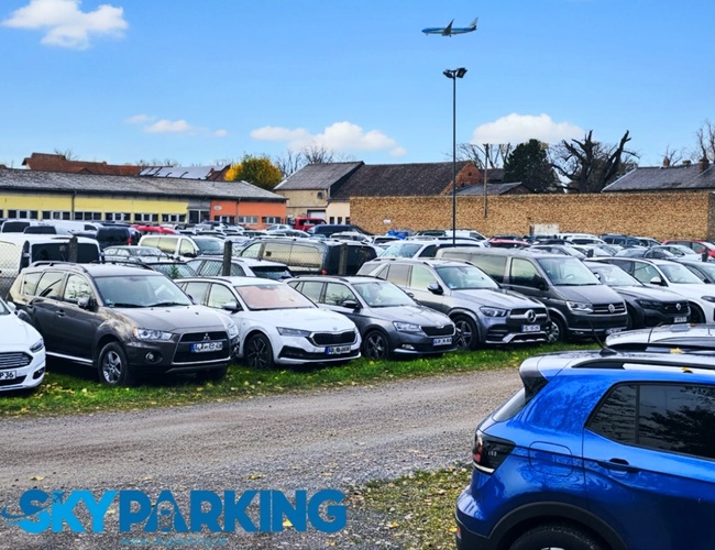 Otwarty parking z wieloma zaparkowanymi samochodami rozmieszczonymi w kilku rzędach, otoczony niską zabudową i oświetlony dziennym światłem.